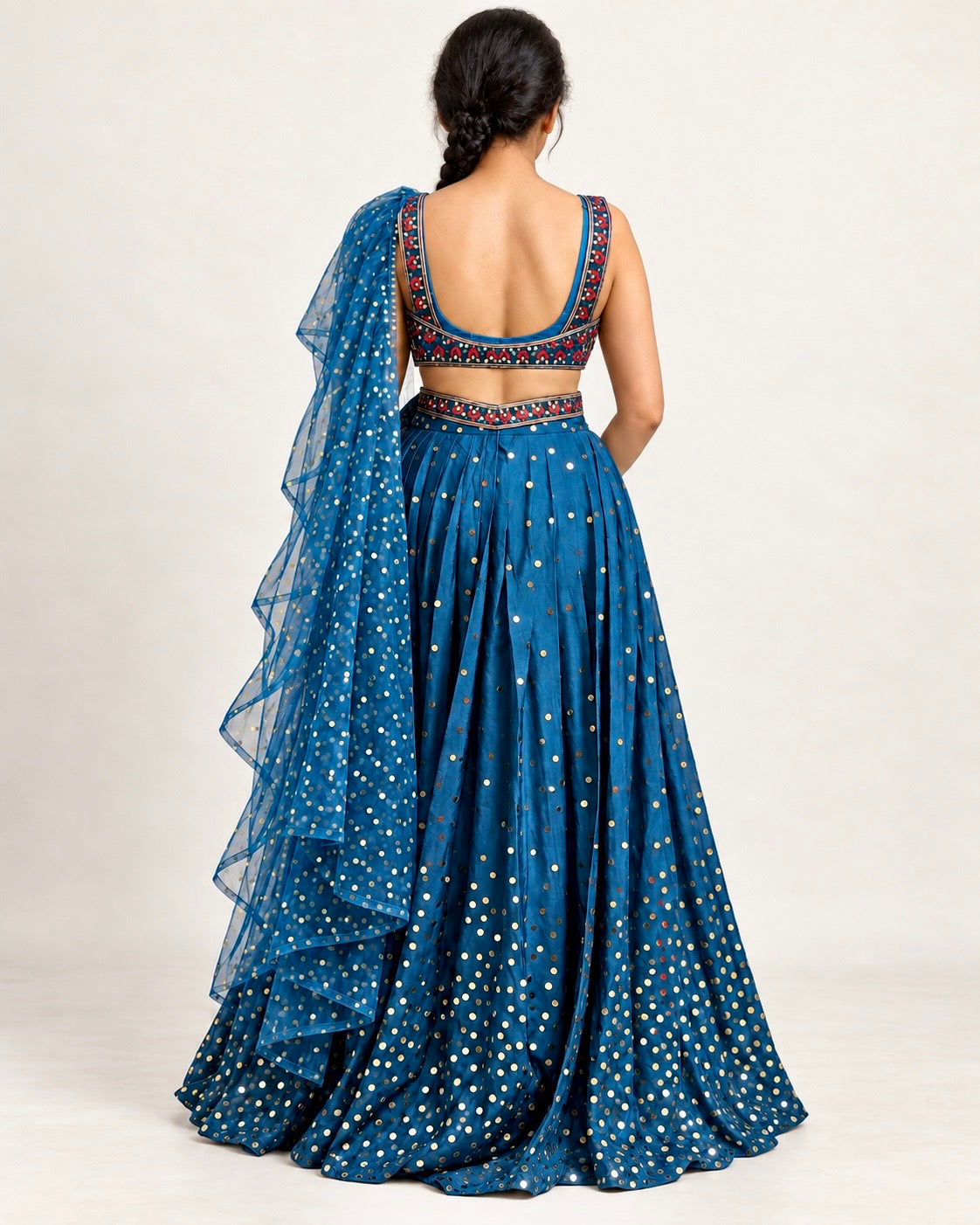 A woman shown from the back wearing a blue traditional lehenga choli set with white printed motifs and embroidered neckline details, photographed against a plain light background.