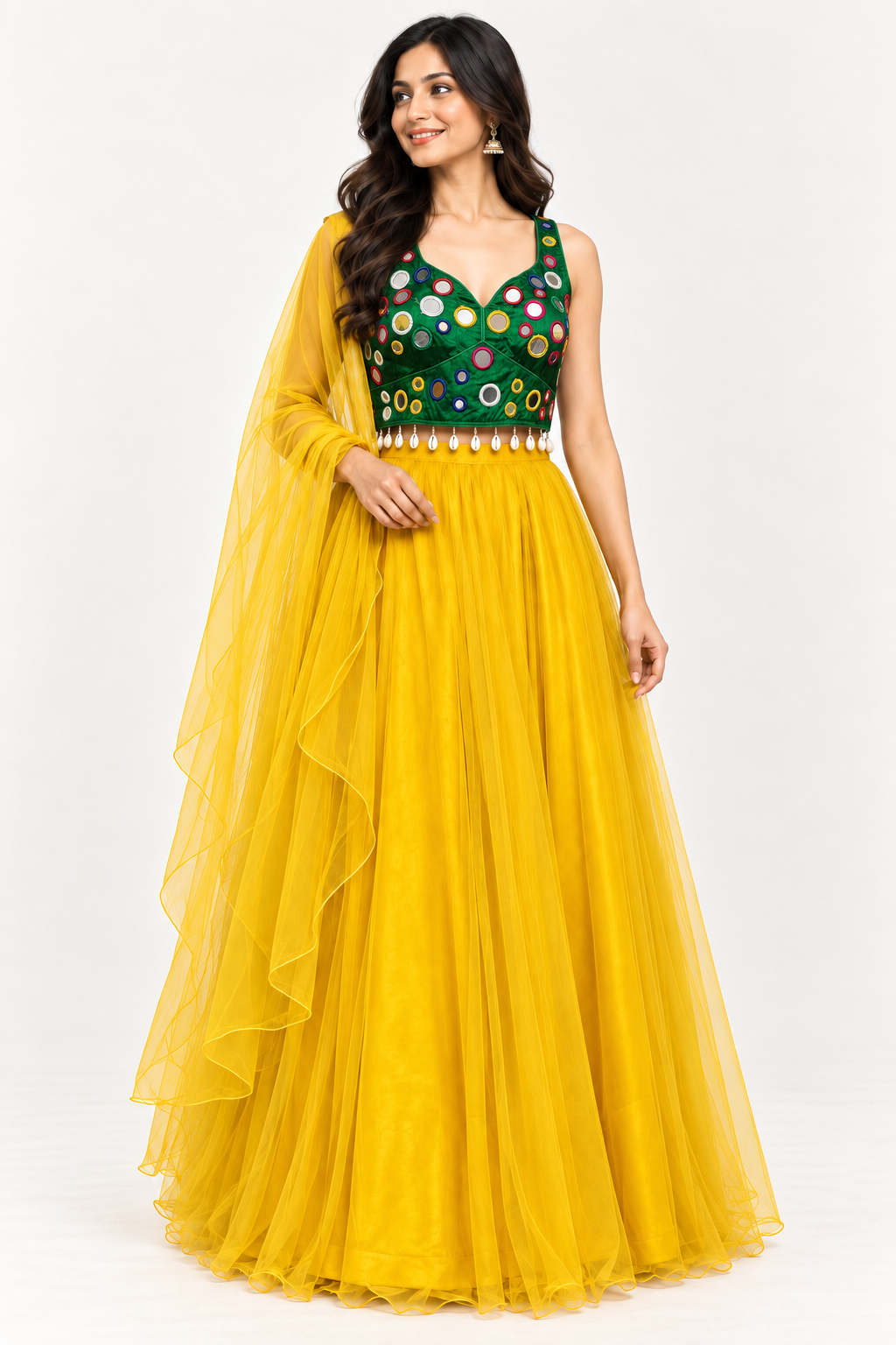 A woman modeling a traditional Indian lehenga choli outfit. The dress features a vibrant yellow tulle skirt and a deep green sleeveless bodice embellished with colorful circular mirror work and gold embroidery, complete with a matching sheer yellow dupatta.