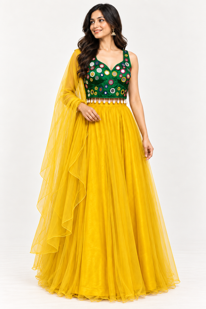 A woman modeling a traditional Indian lehenga choli outfit. The dress features a vibrant yellow tulle skirt and a deep green sleeveless bodice embellished with colorful circular mirror work and gold embroidery, complete with a matching sheer yellow dupatta.
