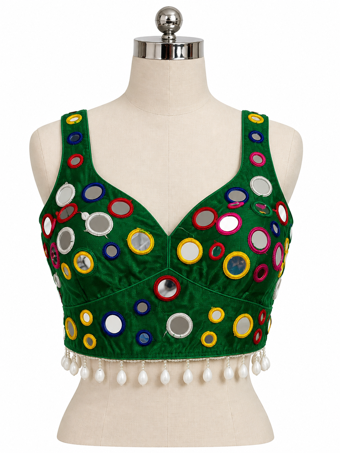 A green sleeveless blouse top with colorful circular mirror work and white tassel fringe, displayed on a cream-colored dress form mannequin against a white background.