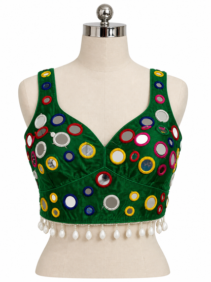 A green sleeveless blouse top with colorful circular mirror work and white tassel fringe, displayed on a cream-colored dress form mannequin against a white background.