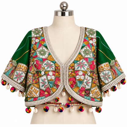 A green embroidered crop jacket with multicolored patchwork embroidery, shell trim, and colorful tassels, displayed on a cream dress form mannequin against a white background.