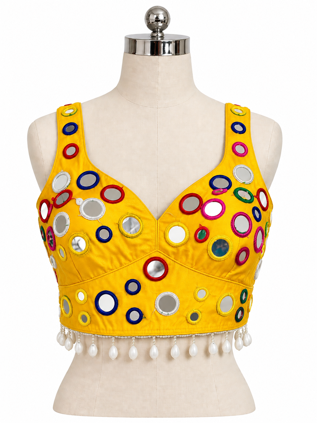 A bright yellow sleeveless crop top blouse displayed on a cream dress form mannequin. The top is decorated with numerous colorful circular mirror embellishments in red, blue, green, and silver, and features a row of white pearl-like tassels along the underbust hem.