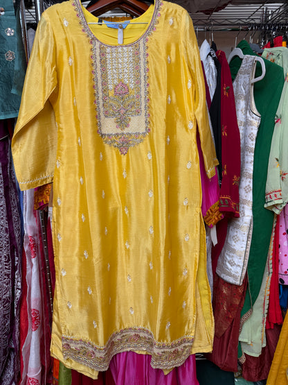 A yellow embroidered kurta hanging on a rack in a clothing store, surrounded by other colorful garments. The kurta features intricate gold and silver embroidery around the neckline and along the hem.